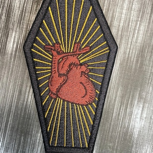 May include: A black and yellow embroidered patch shaped like a coffin with a red anatomical heart inside. The heart is surrounded by yellow rays of light.