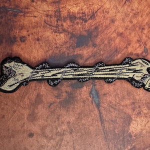 May include: A beige and black embroidered patch of a bone. The bone is detailed with black stitching and has a rough texture.