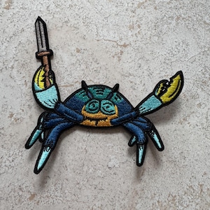 May include: A blue and yellow embroidered crab holding a sword, a whimsical patch for clothing or bags.