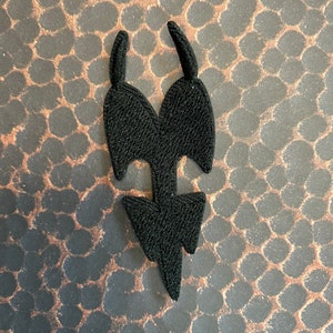 May include: Black fabric patch in the shape of a stylized arrow with two points at the top and a single point at the bottom.