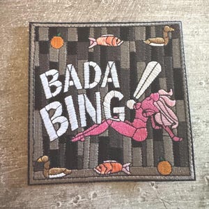 May include: A square embroidered patch featuring a dark gray and black vertical striped background. The text BADA BING is stitched in white. A pink figure, two orange fish, two brown ducks, and two orange circles are embroidered on the patch.