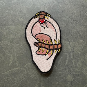 May include: Embroidered patch of a human ear with a centipede crawling inside. The centipede is brown with yellow legs and a red head. The ear is pink and has a detailed outline.