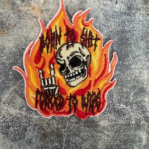 May include: An embroidered patch with a skull in flames. The skull is giving the rock and roll hand sign. The text "Born to Shit" is written around the skull.