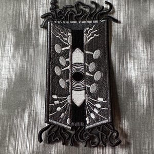 May include: Embroidered patch with a rectangular design. The patch features a central black and white pattern with stylized tree and circle motifs. The border and fringe are black, creating a striking contrast against the silver background.