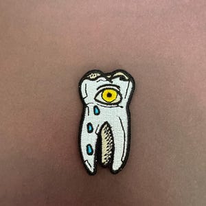 May include: A white tooth with a yellow eye and blue teardrops. The tooth is embroidered on a fabric patch.
