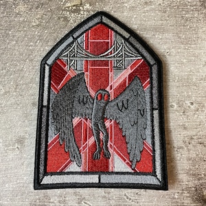 May include: A black and gray embroidered patch featuring a creature with large wings and red eyes, set against a red and white background with a gray bridge.