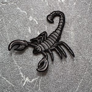 May include: Black embroidered scorpion patch. The scorpion is facing left with its tail curled up. The patch is on a gray textured background.