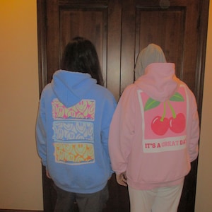 May include: Two people wearing hoodies. The person on the left is wearing a light blue hoodie with a multi-colored smiley face design. The person on the right is wearing a pink hoodie with a white cherry design and the text "It's a great day".