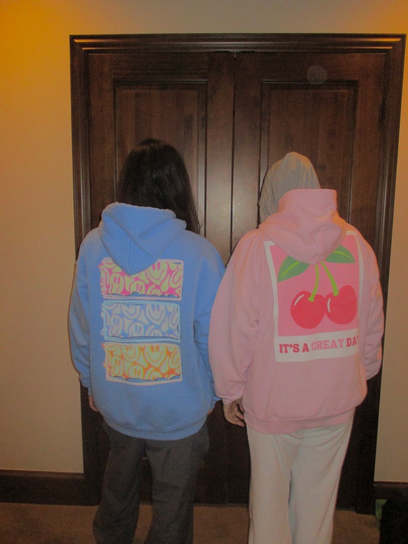 May include: Two people wearing hoodies. The person on the left is wearing a light blue hoodie with a multi-colored smiley face design. The person on the right is wearing a pink hoodie with a white cherry design and the text "It's a great day".