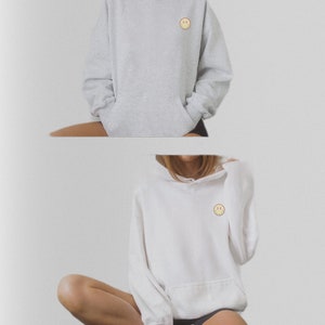 May include: Two hooded sweatshirts with a small yellow smiley face embroidered on the front. One sweatshirt is gray and the other is white.