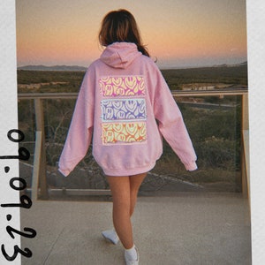 May include: A pink hoodie with a back design of three horizontal stripes featuring a pattern of yellow, purple, and blue smiley faces.