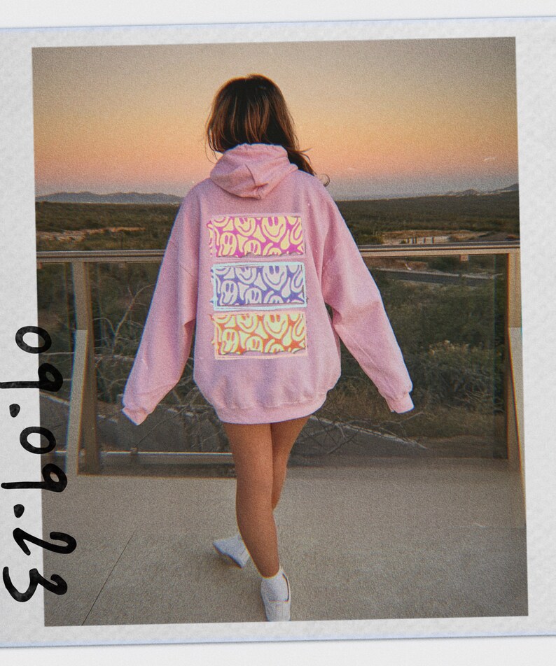 May include: A pink hoodie with a back design of three horizontal stripes featuring a pattern of yellow, purple, and blue smiley faces.