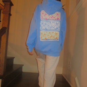 May include: A light blue hoodie with a white pocket on the back. The pocket has a multi-colored smiley face design.