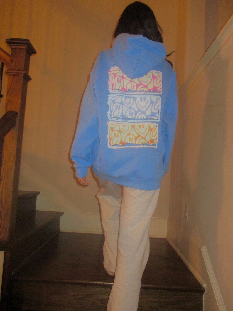 May include: A light blue hoodie with a white pocket on the back. The pocket has a multi-colored smiley face design.