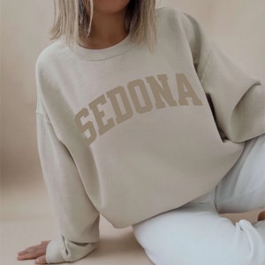 May include: A light beige sweatshirt with the word "SEDONA" printed in a large, brown, arched font.