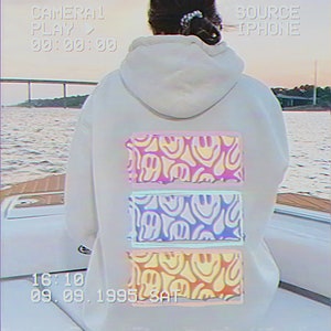 May include: A white hoodie with a pink, blue, and yellow graphic print of smiling faces on the back. The graphic is in a retro style with a faded look. The hoodie has a drawstring hood and a kangaroo pocket.
