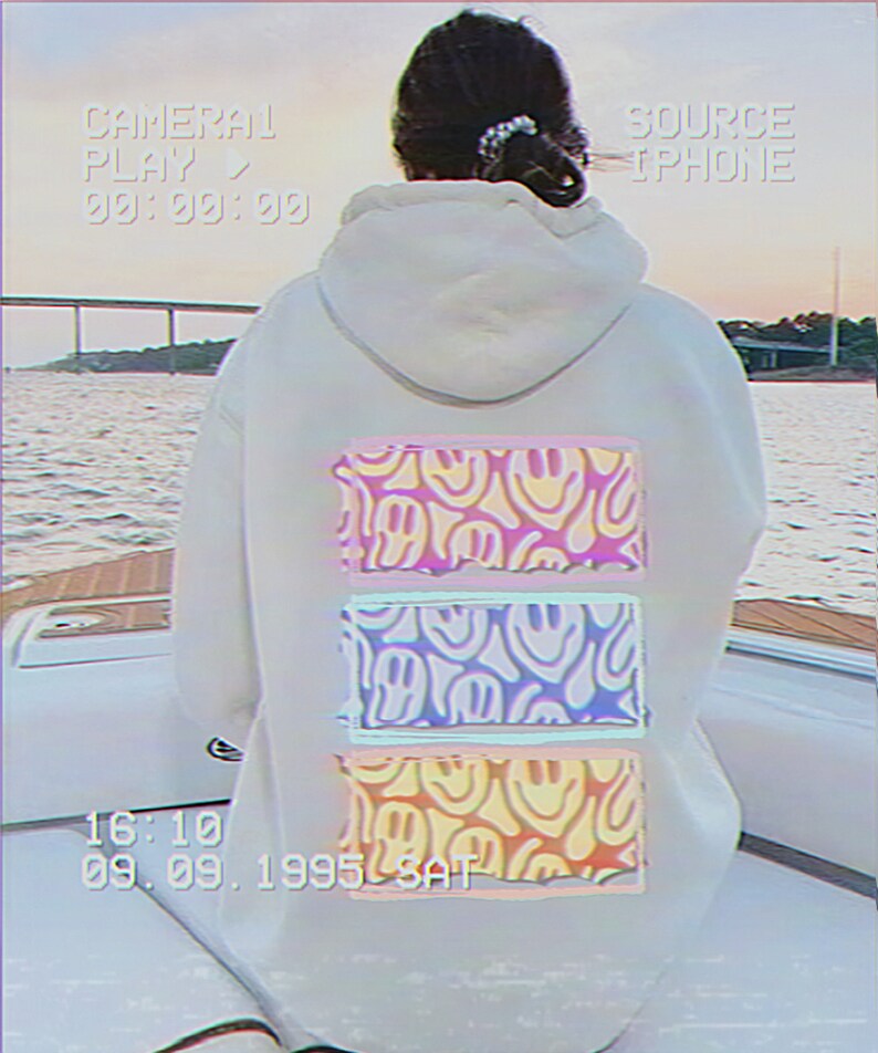 May include: A white hoodie with a pink, blue, and yellow graphic print of smiling faces on the back. The graphic is in a retro style with a faded look. The hoodie has a drawstring hood and a kangaroo pocket.