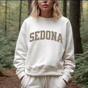 May include: A white sweatshirt with a brown "SEDONA" graphic printed on the front. The sweatshirt has a crew neck and long sleeves.