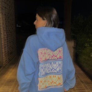 May include: A light blue hoodie with a colorful tie-dye design featuring multiple smiley faces on the back.