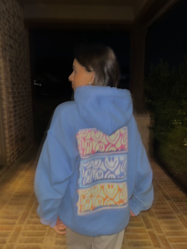 May include: A light blue hoodie with a colorful tie-dye design featuring multiple smiley faces on the back.