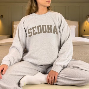 May include: A gray sweatshirt with the word "SEDONA" printed in brown on the front. The sweatshirt is worn by a person sitting on a bed.