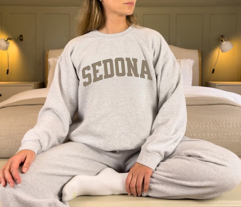 May include: A gray sweatshirt with the word "SEDONA" printed in brown on the front. The sweatshirt is worn by a person sitting on a bed.