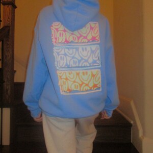 May include: A light blue hoodie with a graphic print of three rows of colorful smiley faces. The hoodie has a hood and a kangaroo pocket.