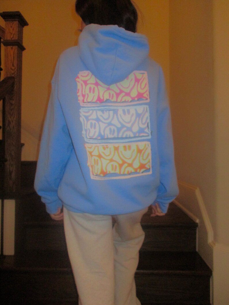 May include: A light blue hoodie with a graphic print of three rows of colorful smiley faces. The hoodie has a hood and a kangaroo pocket.