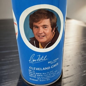 May include: A blue and white Cleveland Cavs collectible cup featuring a portrait of Bill Fitch. The cup includes text with Fitch's name, age, and birthdate. The cup is a vintage sports collectible.