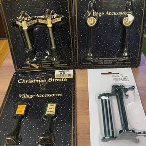 May include: Several Christmas village accessories are displayed in their original packaging. The items include miniature street signs and lampposts, with text like "Main St" and "Ski Lift Ahead." The packaging is black with white stars.