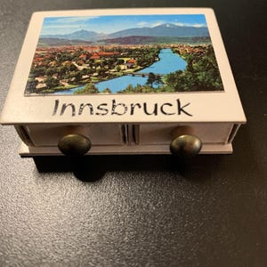 May include: A white wooden box with two drawers and brass knobs. The front of the box has a picture of Innsbruck, Austria, with the word "Innsbruck" printed below the picture.