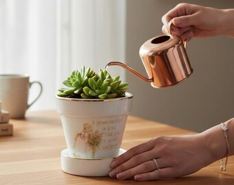 Puede incluir: Una maceta de cerámica blanca con borde dorado y platillo a juego. Contiene una suculenta verde y tiene el texto "A grandmother is love that you never outgrow". Una regadera de cobre riega la planta.
