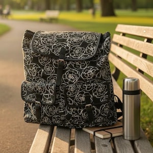 May include: A black backpack with a white floral pattern, two front pockets, and leather straps sits on a wooden bench. A stainless steel thermos and a small notebook are also on the bench, suggesting a day outdoors.
