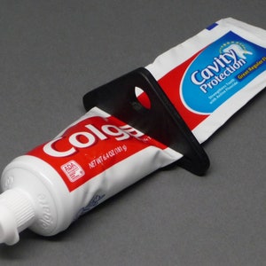 Könnte beinhalten: Eine Tube Colgate-Zahnpasta mit einem schwarzen Kunststoff-Quetscher. Die Tube ist rot und weiß mit dem Text "Colgate" und "NET WT 6.4 oz (181 g)". Ein Päckchen "Cavity Protection" Zahnpasta ist ebenfalls zu sehen.
