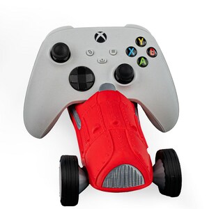 May include: A gray Xbox controller paired with a red toy race car. The controller has black joysticks and colored buttons. The car has black tires and a silver grill.