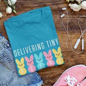 Może przedstawiać: Turkusowa koszulka z napisem "Delivering Tiny Peeps" i pięcioma kolorowymi kreskówkowymi postaciami peeps. Koszulka jest złożona i leży na drewnianej powierzchni z dżinsowymi szortami, różowymi butami sportowymi i białymi słuchawkami.