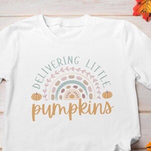 May include: White t-shirt with the text "Delivering Little Pumpkins" in a fall-themed design. The design includes a rainbow, pumpkins, and leaves. The shirt is displayed on a wooden surface with autumn leaves and a pair of blue jeans.