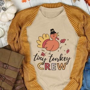 May include: A beige t-shirt featuring a cartoon turkey wearing a pilgrim hat and the text "tiny turkey crew". The shirt is laid out on a wooden surface with a brown cardigan, blue jeans, a pair of sunglasses, a phone case, and autumnal decorations.