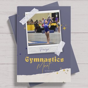 May include: A purple scorebook with a photo of a young gymnast on the cover. The photo is framed with white tape and the text "Paige" is written below the photo. The title of the book is "Gymnastics Meet" in yellow text with a gold star design.