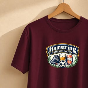Può includere: Una t-shirt a maniche corte color bordeaux con una grafica che riporta il testo "Hamstring AWARENESS SOCIETY". Il design include una scarpa da calcio, un bicchiere di birra, un pallone da calcio e una gamba fasciata, incorniciati da foglie.