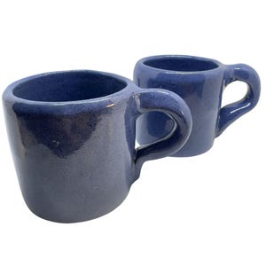 May include: Two handmade ceramic mugs with a glossy, speckled periwinkle blue glaze. Each mug features a thick, rounded wall construction and a substantial, curved handle attached to the side, showcasing a smooth, reflective finish throughout.