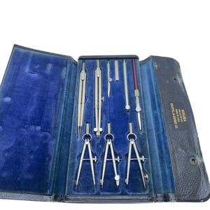 May include: A vintage Keuffel & Esser Minusa drafting instrument set in a black folding case with a blue velvet interior. The set includes various silver-toned metal compasses, dividers, and a drawing pen, all arranged in dedicated slots.