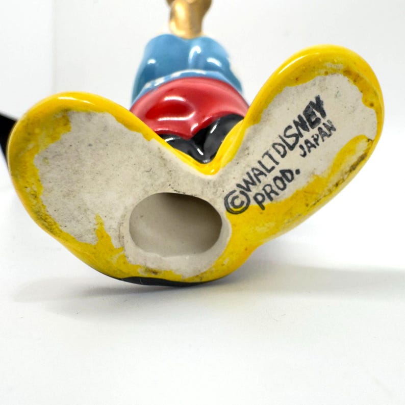 May include: A vintage ceramic figurine of a cartoon character, with yellow feet, red shorts, a blue shirt, and gold accents. The figurine is marked "Walt Disney Prod. Japan."