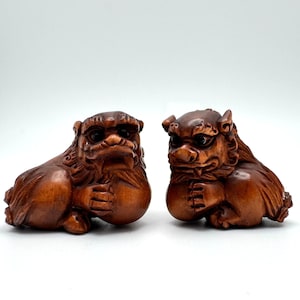 May include: Two wooden sculptures of mythical creatures, possibly Foo Dogs, are depicted. The carvings are a rich brown color, with detailed features including expressive faces and stylized manes. Each figure holds a round object, possibly a pearl, in its paws.