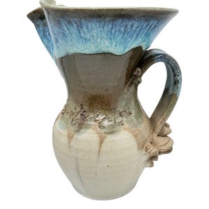Könnte beinhalten: Eine handgefertigte Keramikkrug mit einem einzigartigen Design. Der Krug hat einen hellblauen Rand, der in Braun- und Cremetöne übergeht. Der Griff ist mit einem floralen Akzent versehen.