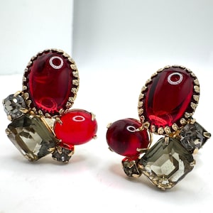 May include: A pair of vintage clip-on earrings. Each earring features a large, oval, ruby-red glass stone surrounded by a gold-tone metal frame. Smaller red and gray stones are also included in the design.
