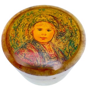 May include: A circular wooden trinket box featuring a painted portrait of a young girl on the lid. The portrait shows a girl with blonde hair and a colourful headscarf, set against a yellow and green background. The box has a light brown exterior.