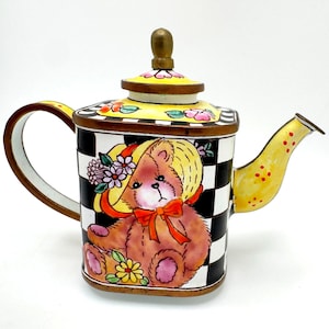 May include: A square ceramic teapot featuring a black and white checkered pattern. The front displays a brown teddy bear wearing a yellow hat with flowers and an orange bow. The lid is yellow with pink flowers and a brown knob, and the spout is yellow.