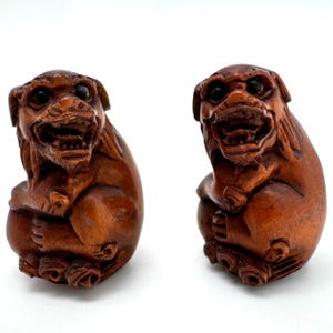 May include: Two intricately carved wooden sculptures of mythical creatures. Each figure is seated, with a fierce expression, open mouth, and detailed fur. The sculptures are a warm brown color and appear to be hand-carved.