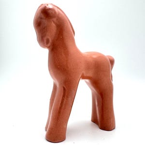Puede incluir: Figura de caballo de cerámica color terracota. El caballo está estilizado con una forma suave y redondeada y un cuello ligeramente curvado. La figura tiene un acabado brillante y se apoya sobre cuatro patas.